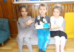 Troje dzieci siedzi na kanapie. W środku chłopiec trzyma pracę: pingwiny zrobione z rolek po papierze stojące na krze z tacki po jedzeniu i korków. Po bokach siedzą dziewczynki i dłońmi pokazują na wykonaną wspólnie pracę.